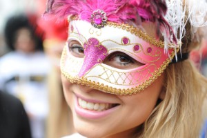Una imagen de una mujer rubia sonriente que lleva una máscara rosa, blanca y dorada con plumas rosas sobre la nariz, los ojos y la frente.